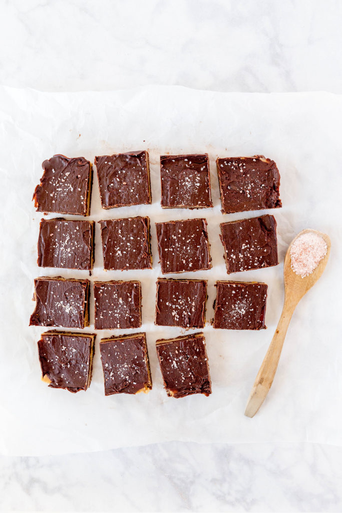 raw caramel slice sitting on parchment paper with a wooden spoon containing sea salt flakes