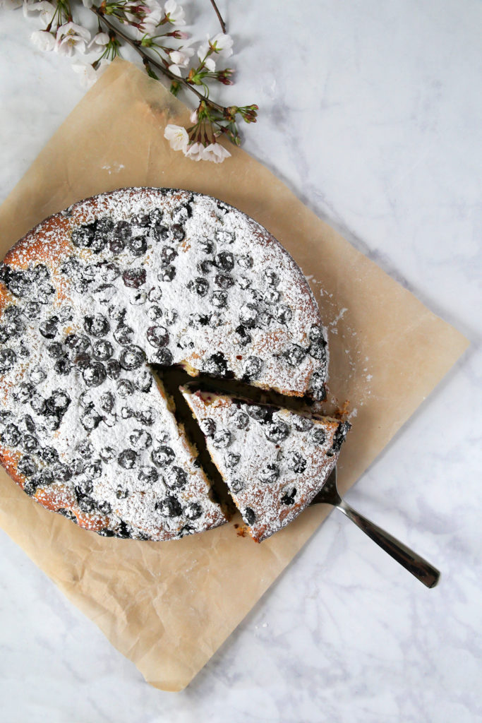 Blueberry cake sitting on brown parchment paper with one slice cut