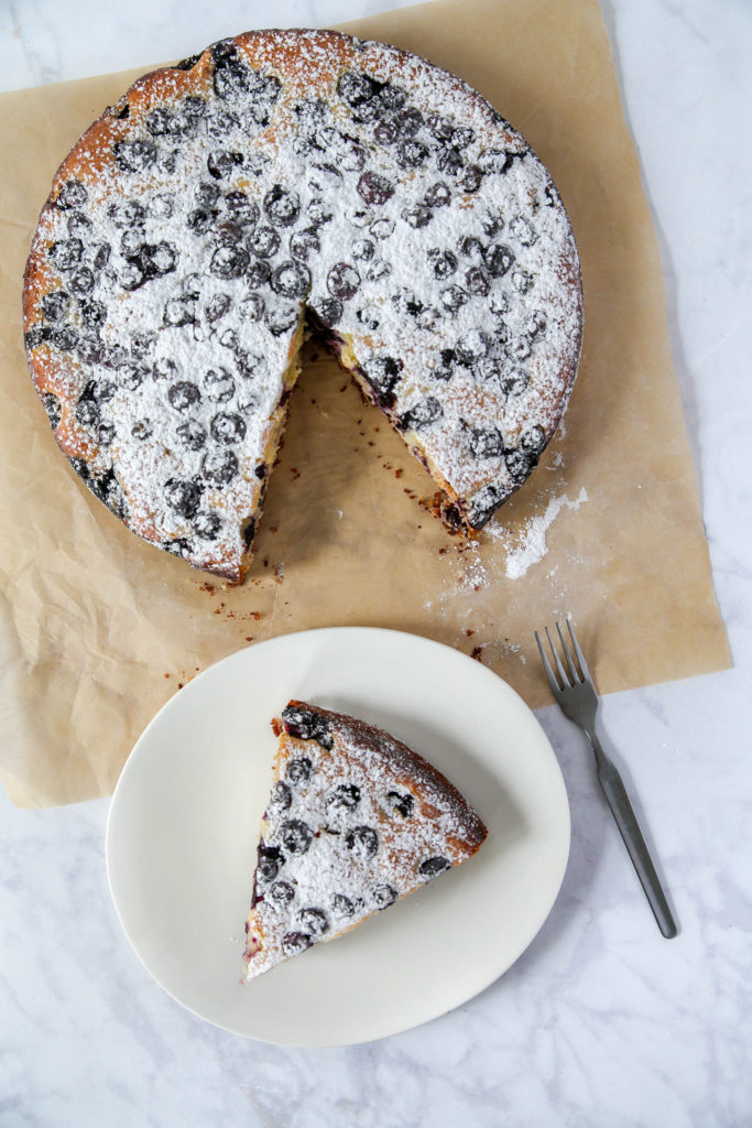 A whole blueberry lemon cake with one slice cut out, sitting on a white plate