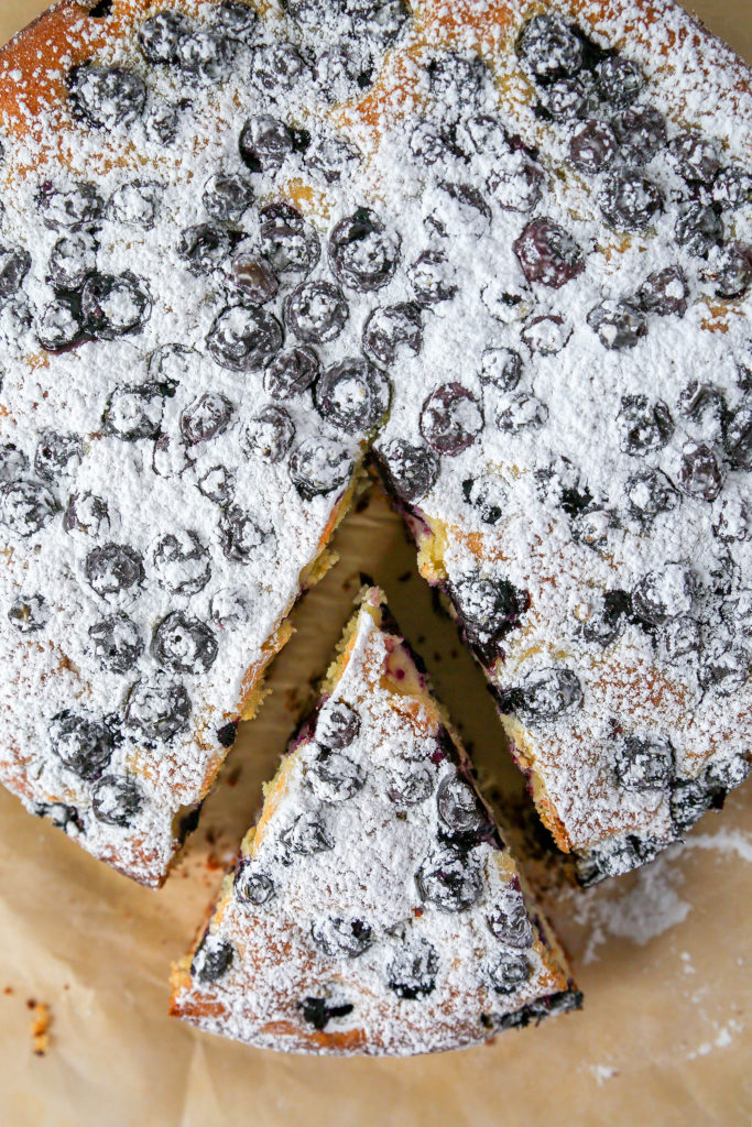 Blueberry lemon cake with one slice cut, sitting on brown parchment paper