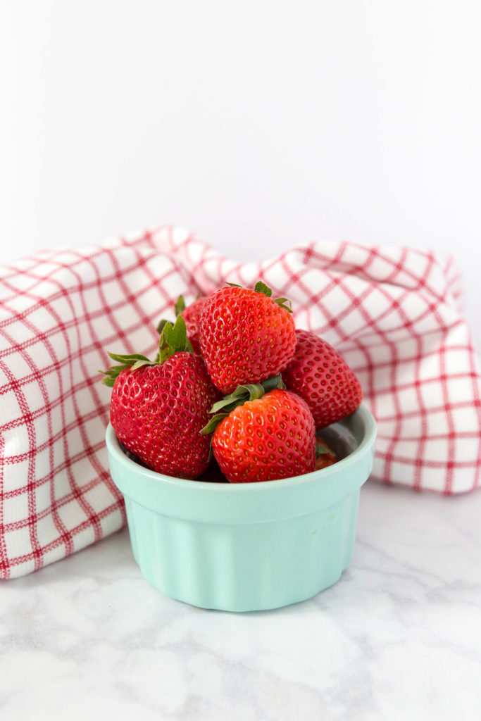An aqua coloured ramekin filled with fresh strawberries, with a red check dish towel behind it. 