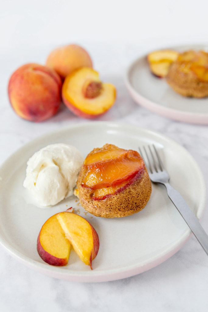 Mini peach upside down cake on a white plate with a scoop of vanilla ice cream and two slices of peach, with 3 peaches in the background 