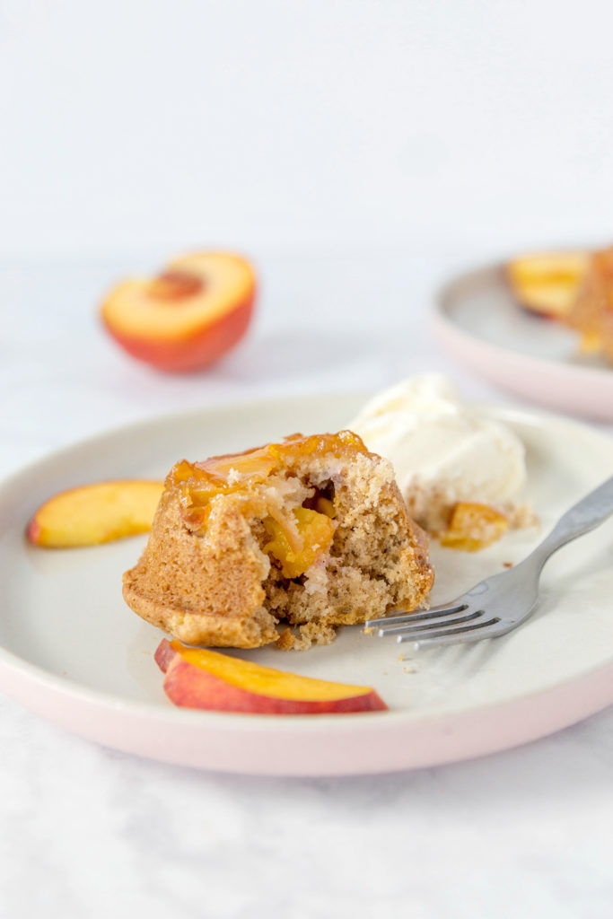 Half of a mini peach cake on a plate to show the inside. Peach slices and vanilla ice cream on the plate.