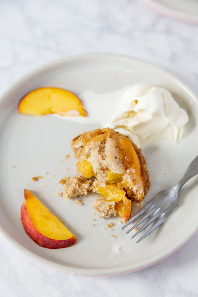A half-eaten peach mini cake to show the inside. Vanilla ice cream and 2 peach slices are also on the plate.