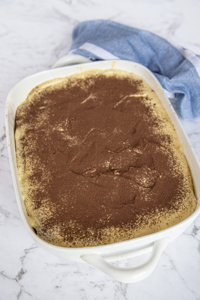 Tiramisu dessert in a white pan, sitting on a marble counter, with a blue kitchen towel at the back of the photo