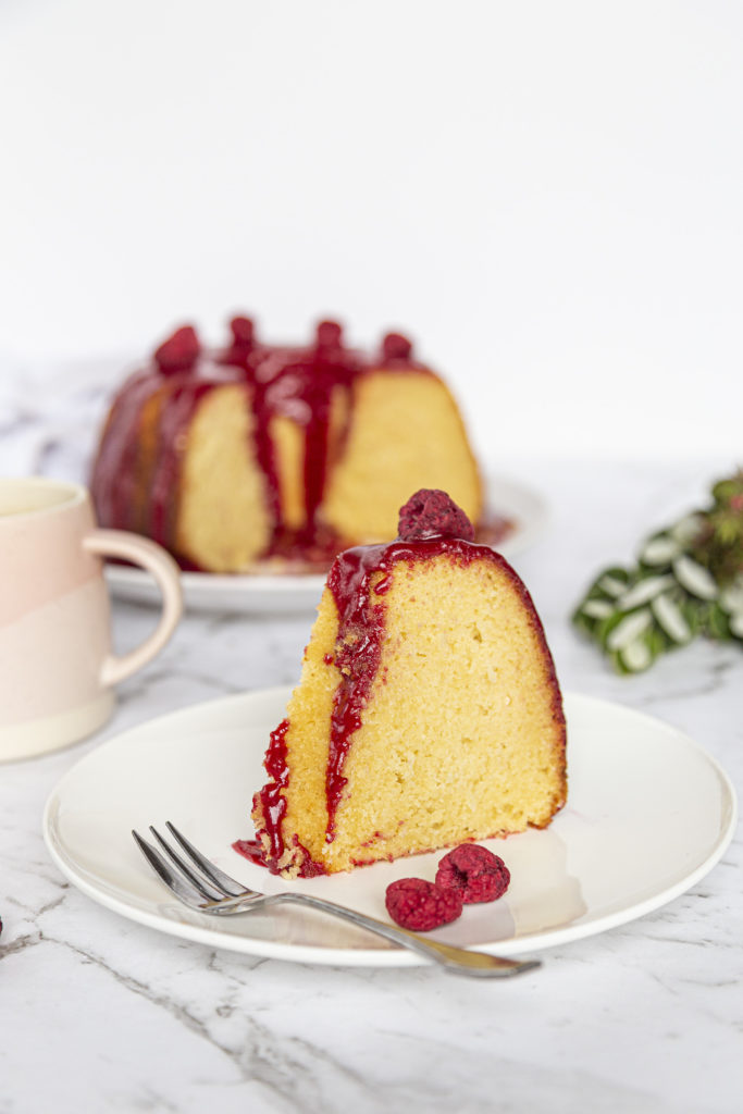 A slice of coconut raspberry bundt cake sitting on a white plate with a small cake fork. The rest of the cake is in the background, along with a pink coffee mug to the left and a green plant to the right.