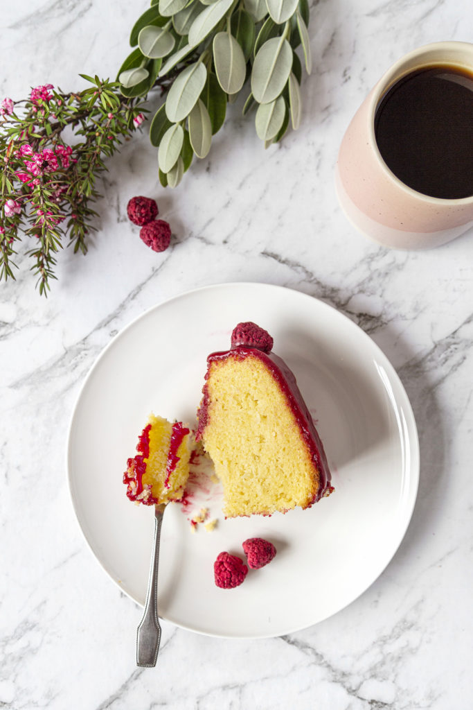 A slice of coconut raspberry bundt cake sitting on a white plate with a small fork. A cup of coffee in a pink mug next to the plate. And a green and pink plant above.