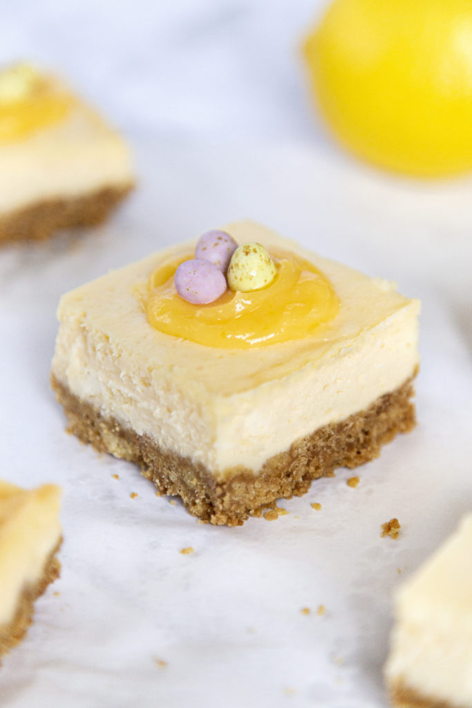 A lemon cheesecake bar topped with lemon curd and mini chocolate eggs, with a lemon in the background