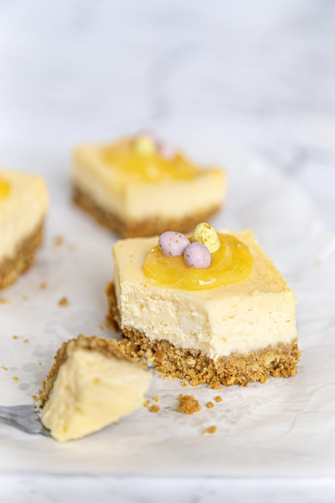 A lemon cheesecake bar topped with lemon curd and mini chocolate eggs, with a bite taken out resting on a fork.