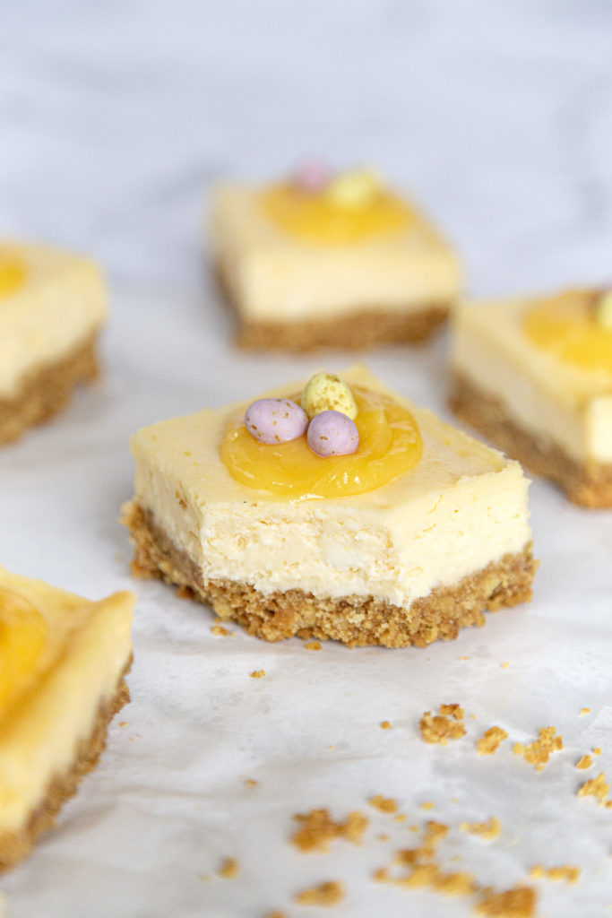 Several lemon cheesecake bars topped with lemon curd and mini chocolate eggs with a bite taken out of one.