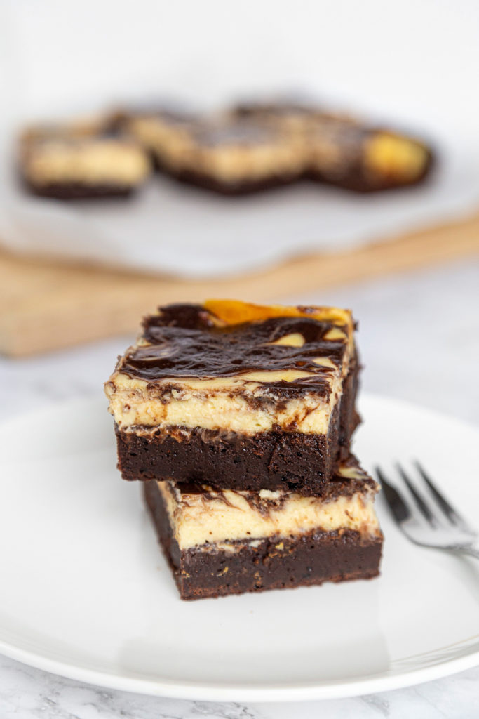 Two cheesecake brownies stacked on a white plate, with a fork