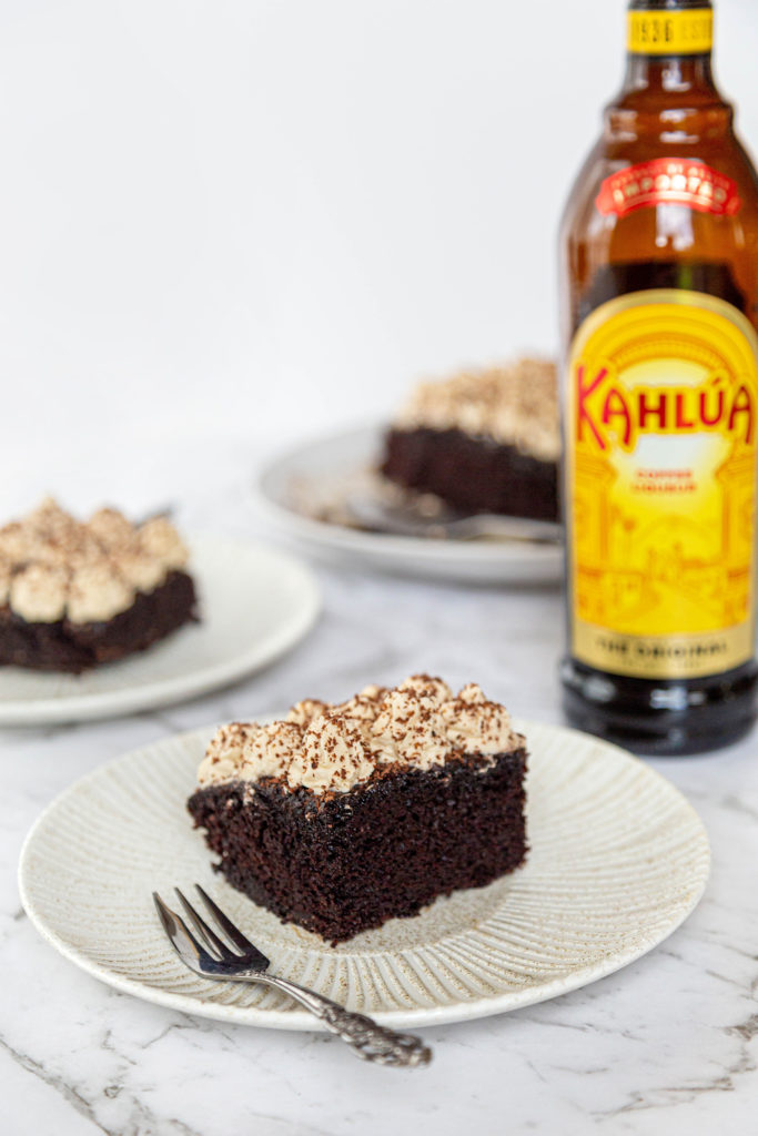 A slice of kahlua chocolate cake on a cream plate with a small fork. A bottle of Kahlua behind the cake.