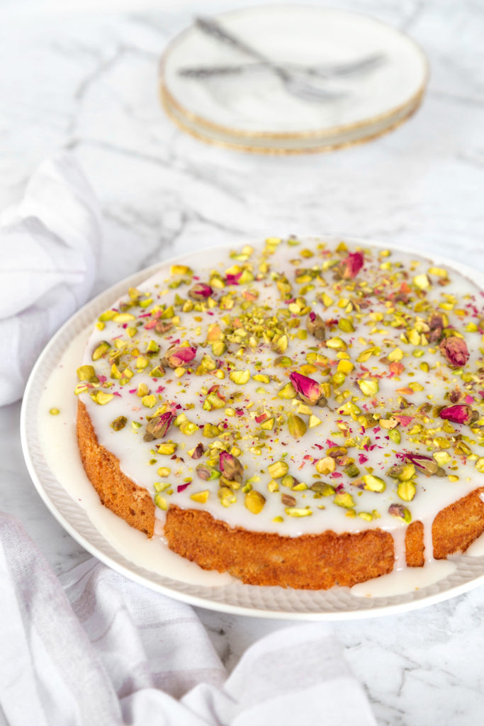 Persian Love cake with white glaze, chopped pistachios and rose petals, sitting on a white plate.