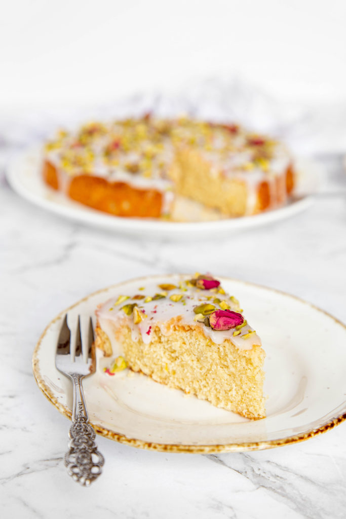 A slice of Persian Love cake with white glaze, chopped pistachios and rose petals, sitting on a white plate with small silver fork.