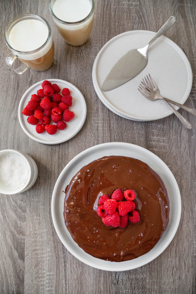 Flat-lay of a whole chocolate cake sitting on a white plate with a pile of raspberries in the center of the cake. Serving plates, forks and coffee surround the cake.