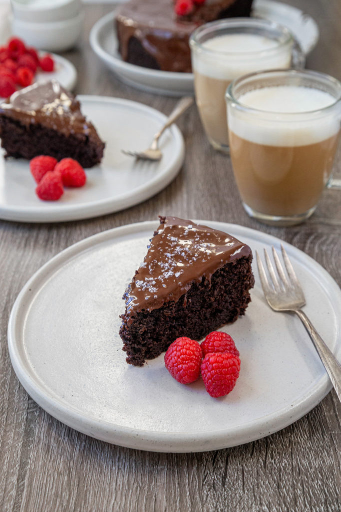 A slice of chocolate cake sitting on a white plate with raspberries on the side. A cup of coffee sitting behind the cake.