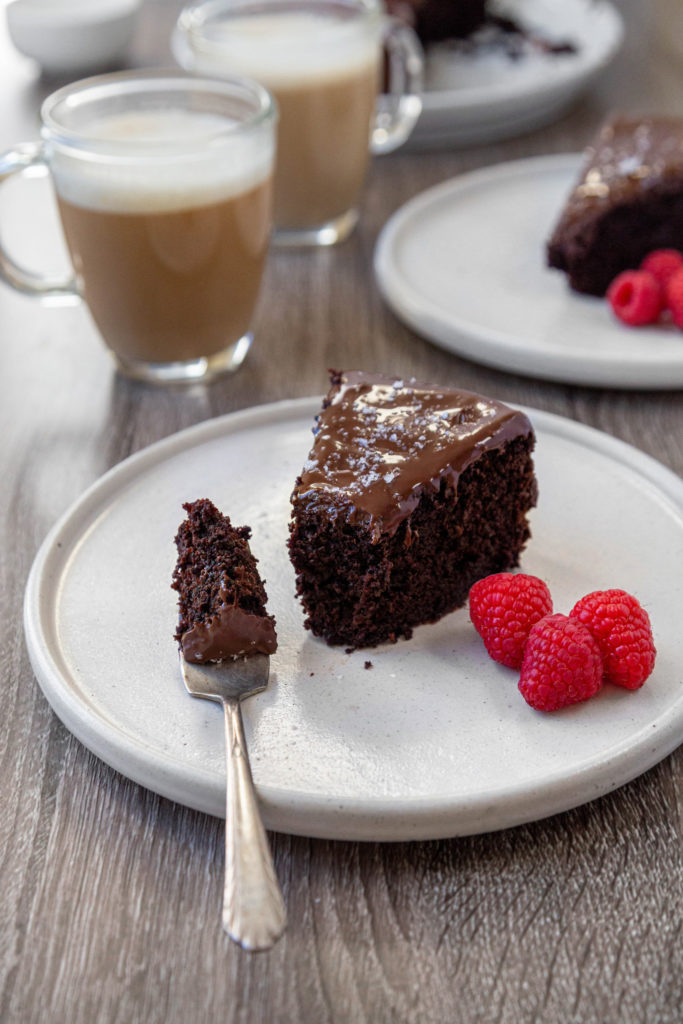 A slice of chocolate cake with a mouthful sitting on a fork on a white plate with raspberries