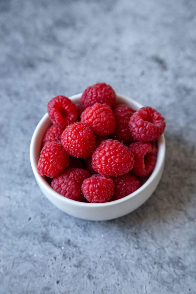 A white bowl with fresh raspberries sitting on a blue/grey background