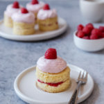 Mini raspberry cake siting on a white plate with a fork. A bowl of raspberries are in the background, as well as a plate of cakes and a coffee cup.