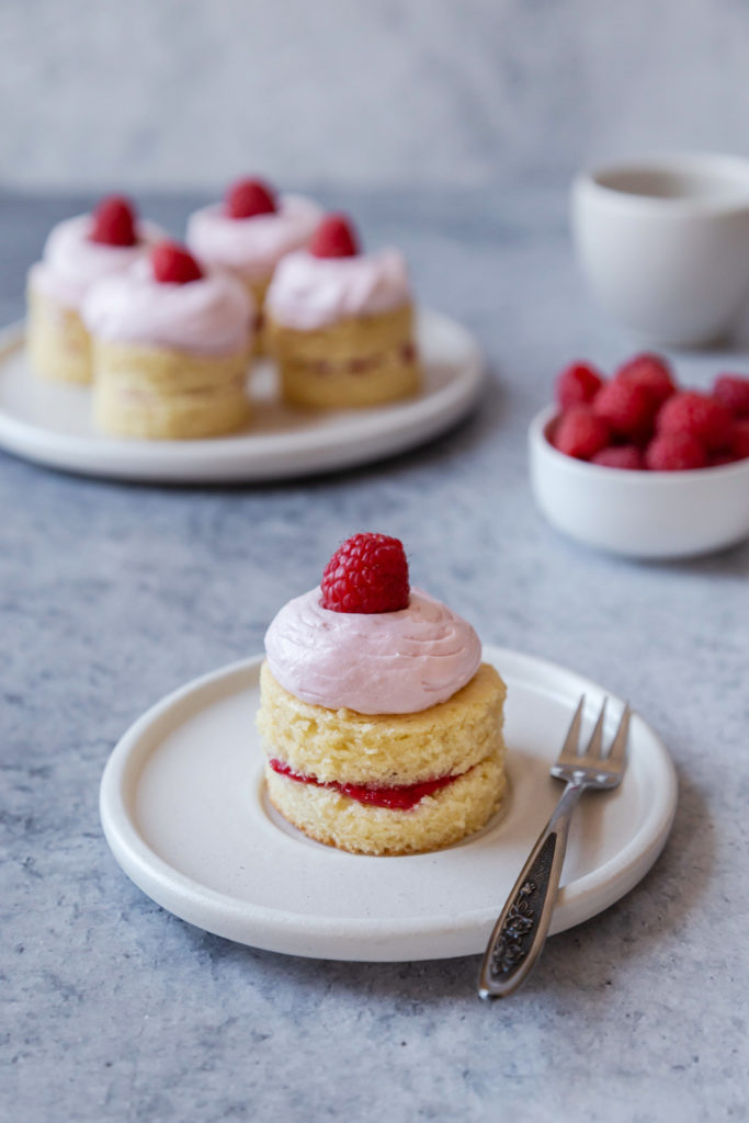A mini raspberry cake sitting on a white plate with a fork. A bowl of raspberries in the background, with a plate of mini raspberry cakes next to it. 