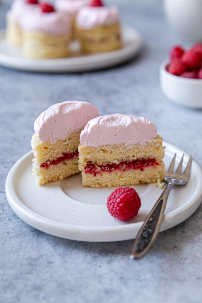 Cross-section of a mini raspberry cake, sitting on a white plate with silver fork. A bowl of raspberries is in the background, as well as a plate of mini cakes.  