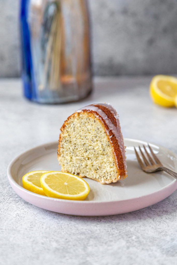 A slice of lemon poppy seed cake sitting on a pink-rimmed plate with a fork and a slice of lemon