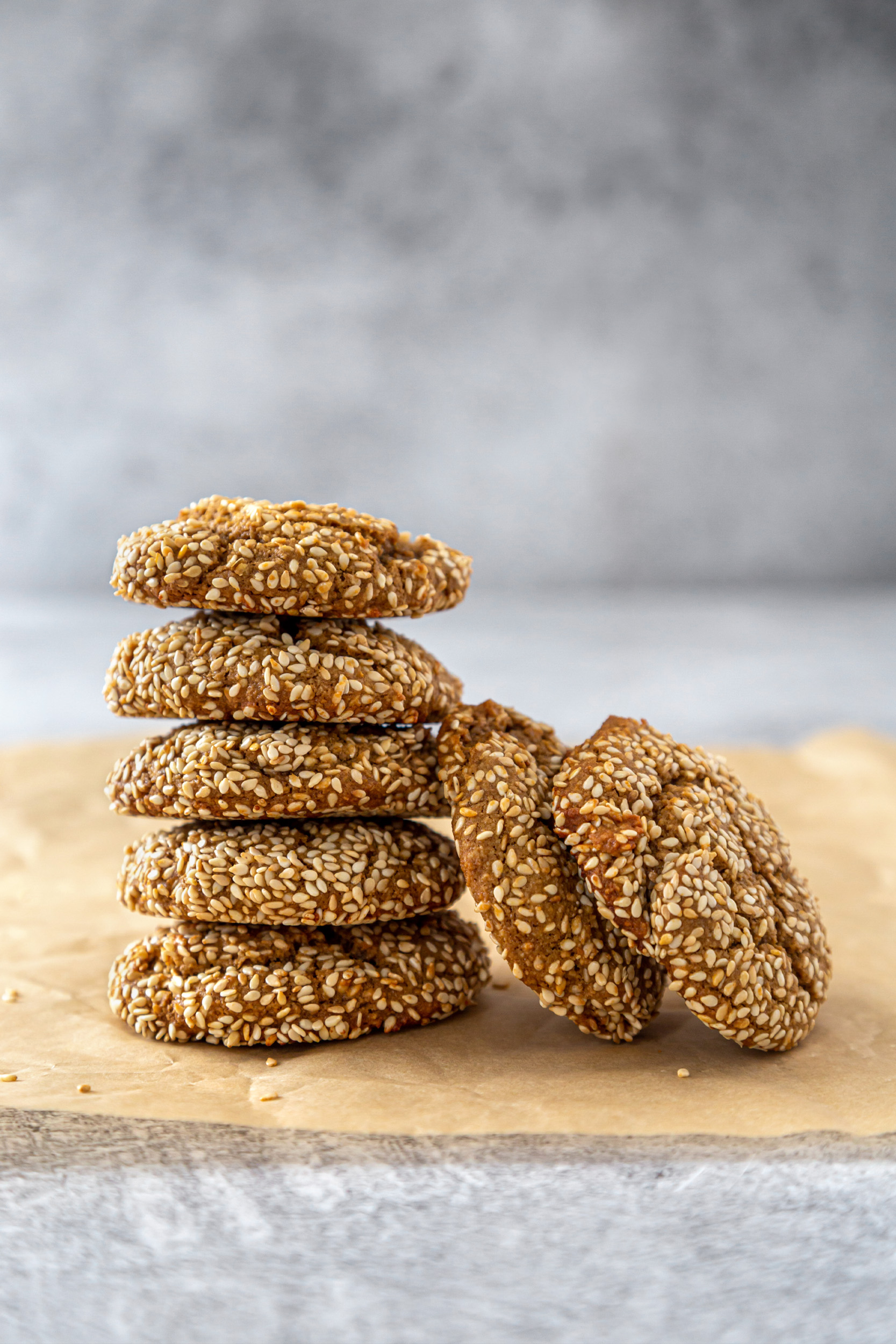5 sesame tahini cookies stacked in a tower, with 2 other cookies leaning against it, on brown parchment paper