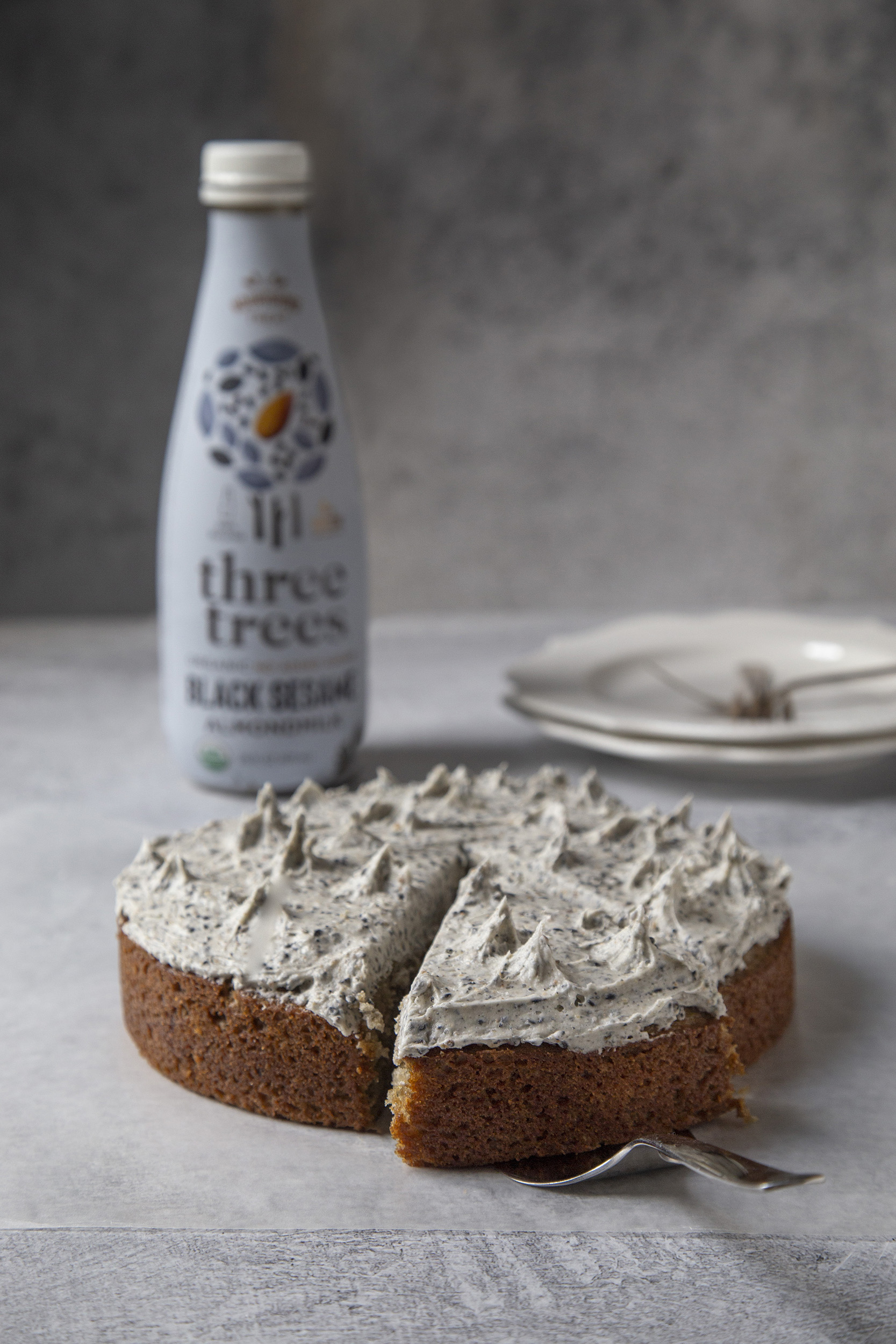 Photo of black sesame cake in the foreground with one slice cut. In the background is white plates and forks, and black sesame almond milk bottle
