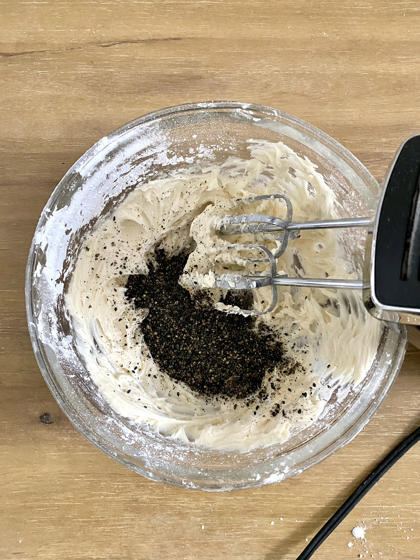 black sesame seeds being added to frosting in glass bowl with hand mixer to the side