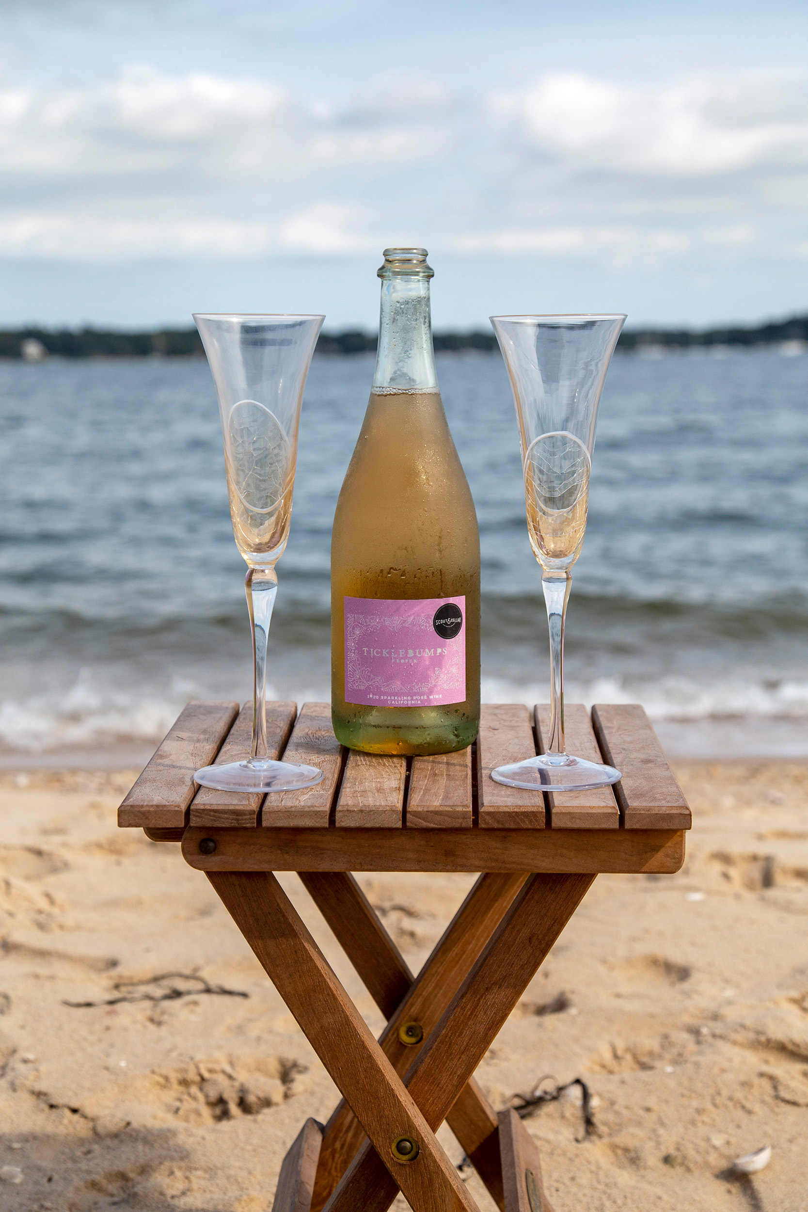 Two flute glasses on a small wooden table, with a bottle of wine in the middle. On a beach with the sea in the background