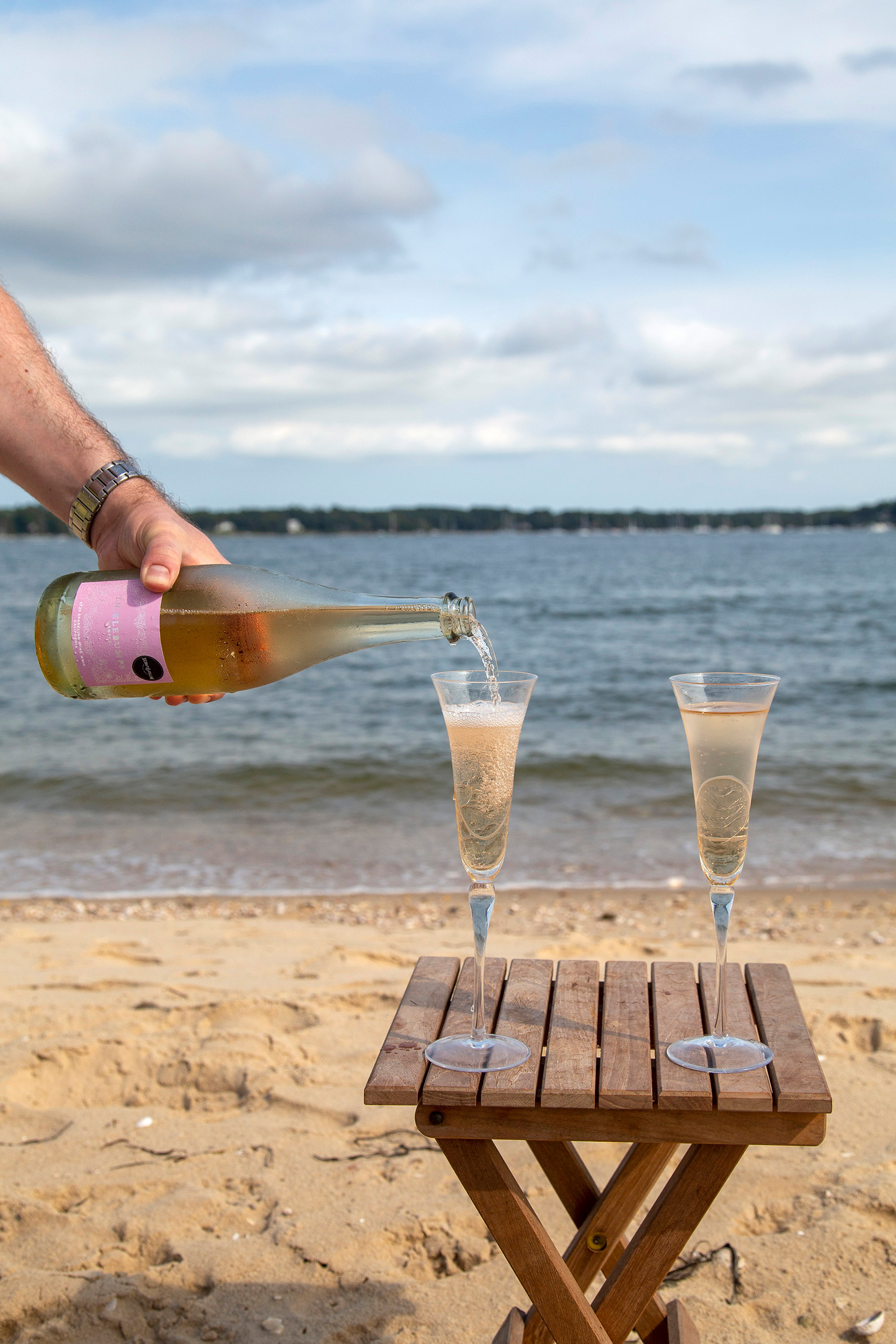 Two flute glasses on a small wooden table, with a bottle of wine being poured at the left side. On a beach with the sea in the background