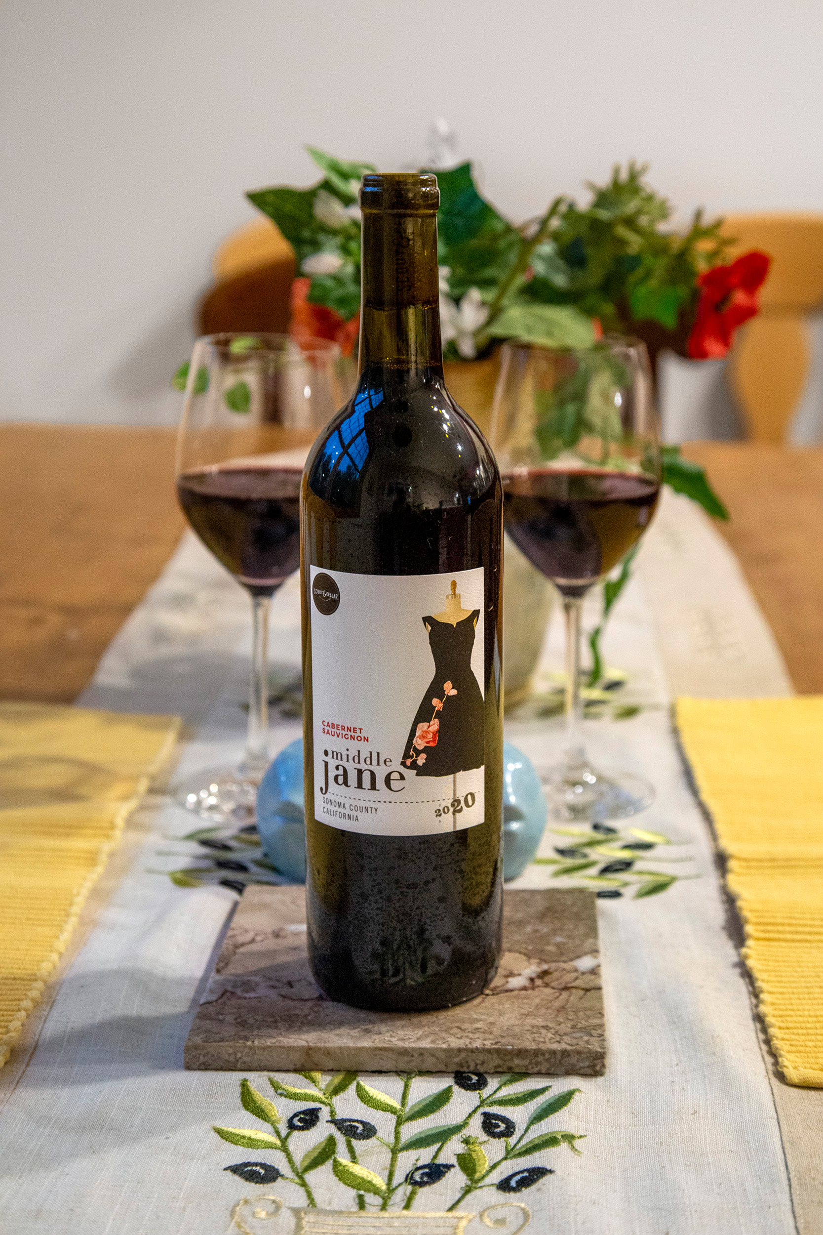 A bottle of red wine sits on a dining room table with two wine glasses in the background