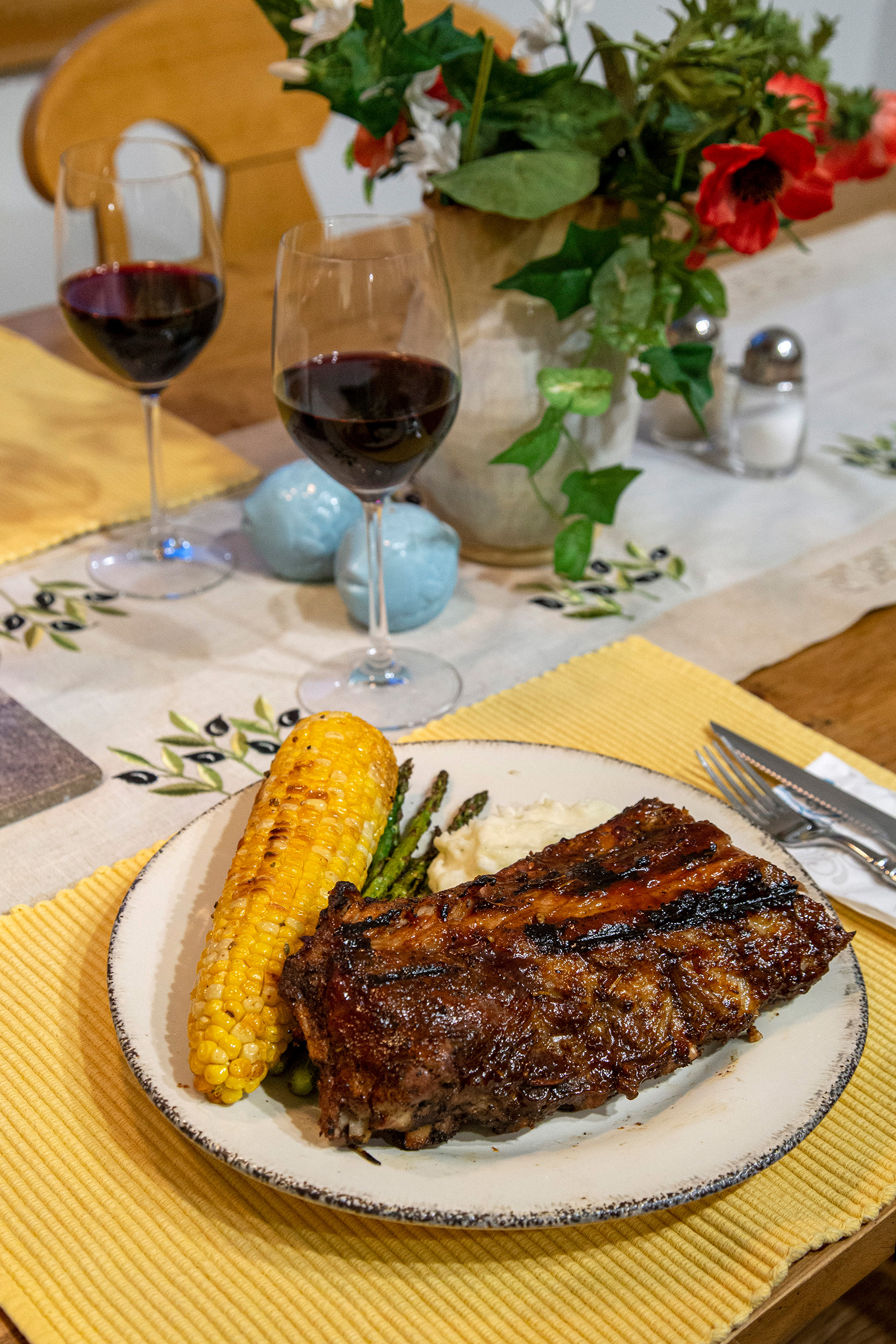 Two wine glasses sit on a dining room table with a plate of bbq ribs and corn in the foreground, with yellow placemats