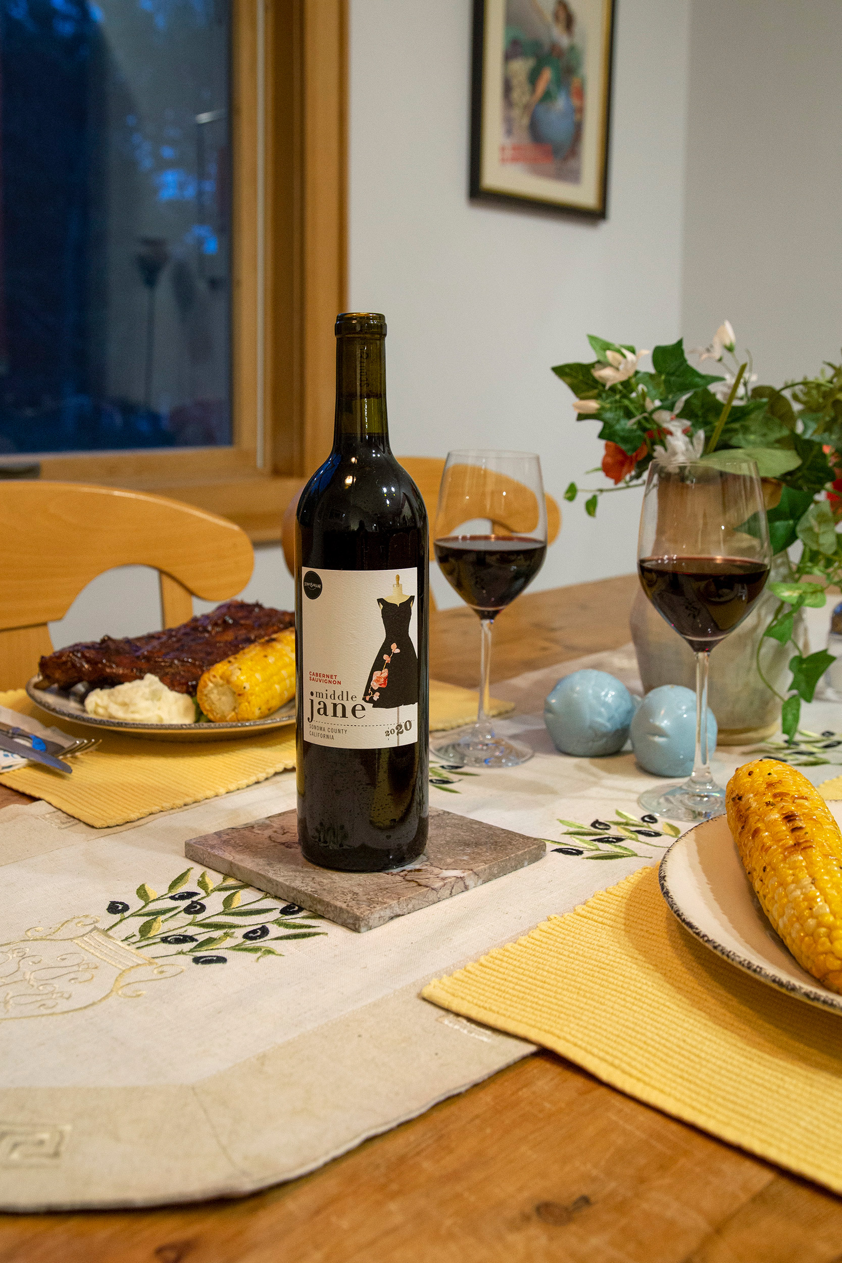 A bottle of red wine sits on a dining room table with dinner plates and two wine glasses