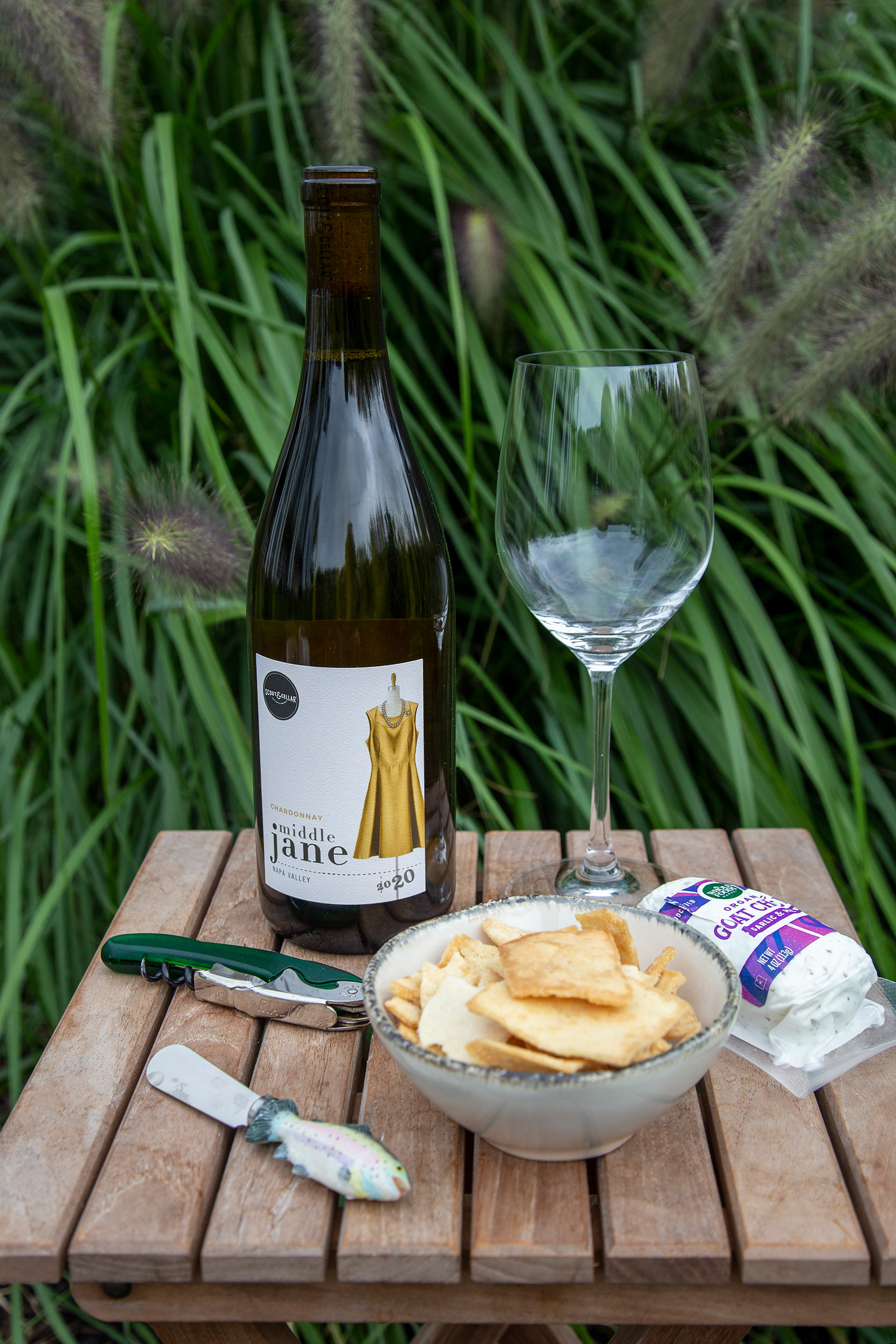 A bottle of wine sitting on a small wooden table with a wine glass, goat cheese, a bottle opener, a cheese knife and a small bowl of pita chips